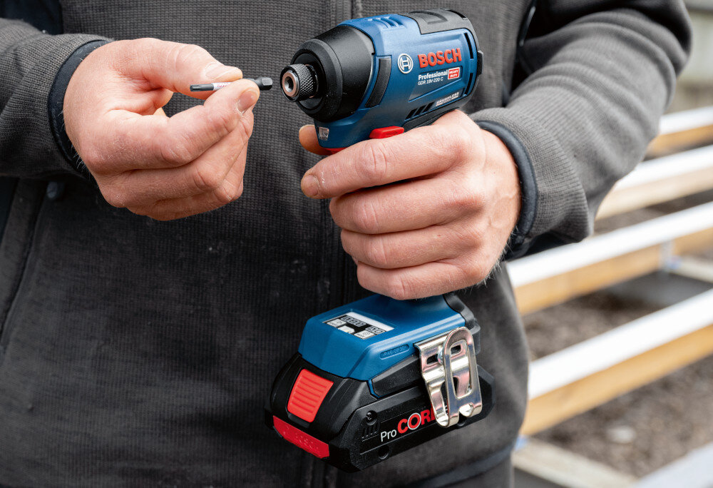 Klucz udarowy BOSCH Professional GDR 18V-220 C 06019L6000 podwórko osoba trzyma klucz w dłoni w drugiej ma bit przystawia go do uchwytu w tle drewniany podest sześciokątny uchwyt 1/4 cala współpracuje ze standardowymi bitami blokowanie jednym ruchem dłoni skraca to czas wymiany osprzętu przy częstych zmianach końcówek