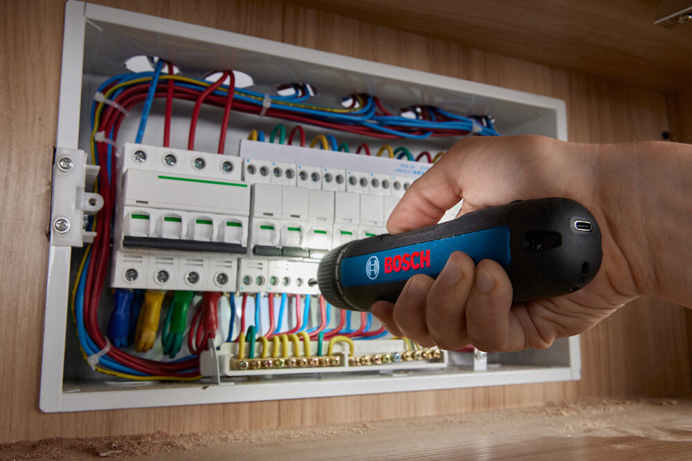Wkrętarka akumulatorowa BOSCH GO Professional Gen III 06019H2201 3.6V 2Ah ściana skrzynka z kablami i bezpiecznikami osoba używa wkrętarki zintegrowany pierścień LED precyzja w słabo oświetlonych przestrzeniach organizacja zadań płynność realizacji projektów