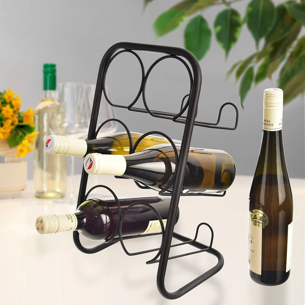 Czarny metalowy stojak na wino mieszczący sześć butelek, z trzema butelkami wina umieszczonymi poziomo. W tle widoczne są inne butelki oraz zielone liście rośliny.
