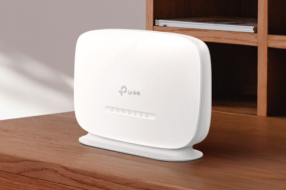 Router TP-LINK TL-MR105 Biały router z logo TP-Link, ustawiony na drewnianej powierzchni w pobliżu półki z książką. Na przednim panelu znajduje się rząd małych diod LED, opis
