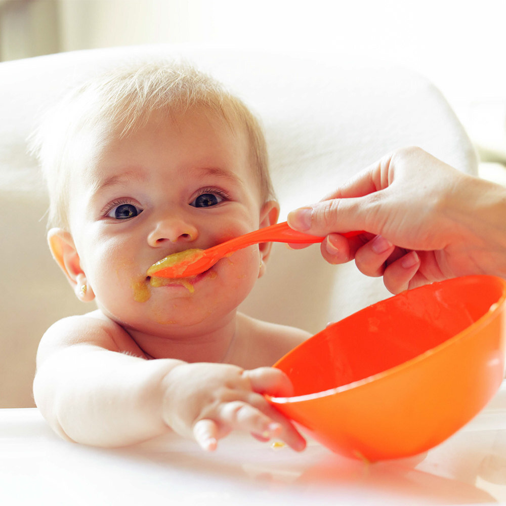 Dziecko jest karmione pomarańczową łyżeczką z pomarańczowej miseczki. Na twarzy dziecka widać resztki jedzenia.