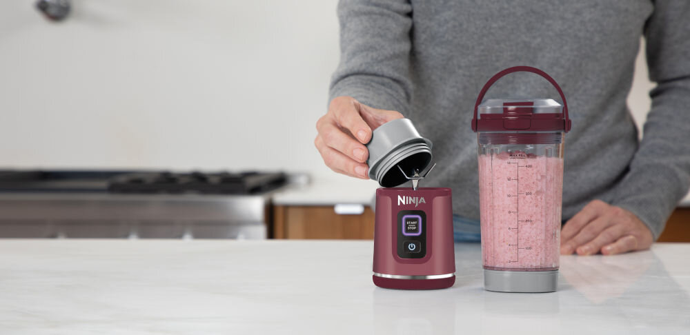 Ninja BC151EUCR blender przenośny stoi na blacie pojemnik z napojem wewnątrz osoba otwiera podstawę blendera z ostrzami przycisk na obudowie kubek pokrywka wolne od BPA dla zdrowego i wygodnego gotowania możliwość mycia w zmywarce funkcja samoczyszczenia woda kropla płynu do naczyń 30 sekundowy cykl miksowania płukanie dla bezpieczeństwo ostrzy zakładanie pokrywy ochronne