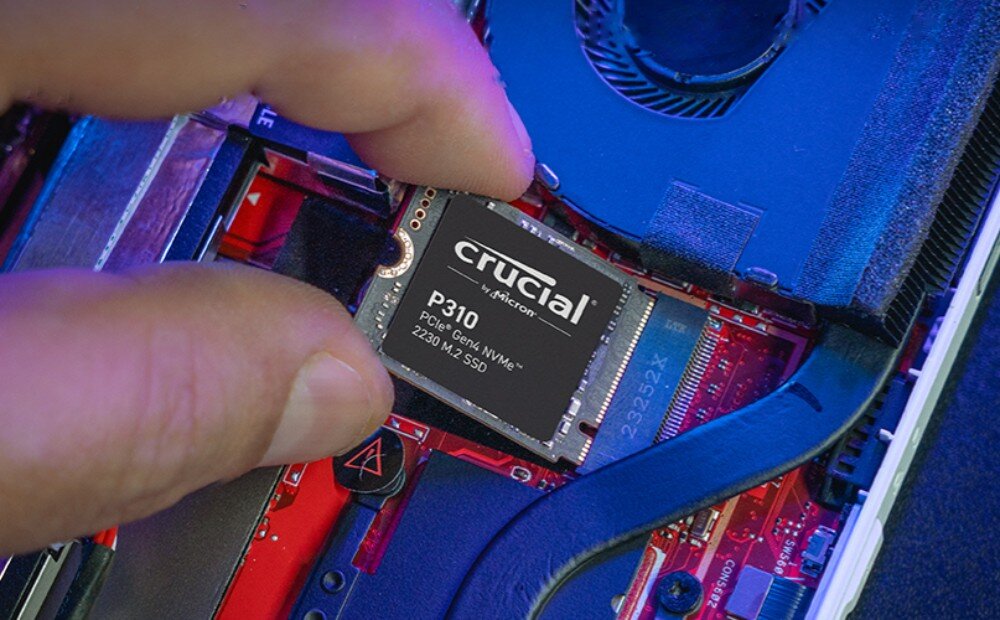 Dysk CRUCIAL P310 Dłoń montująca niewielki nośnik danych w gnieździe płyty głównej przenośnej konsoli lub laptopa, z widocznym czerwonym laminatem i chłodzeniem, interfejs, szybkość