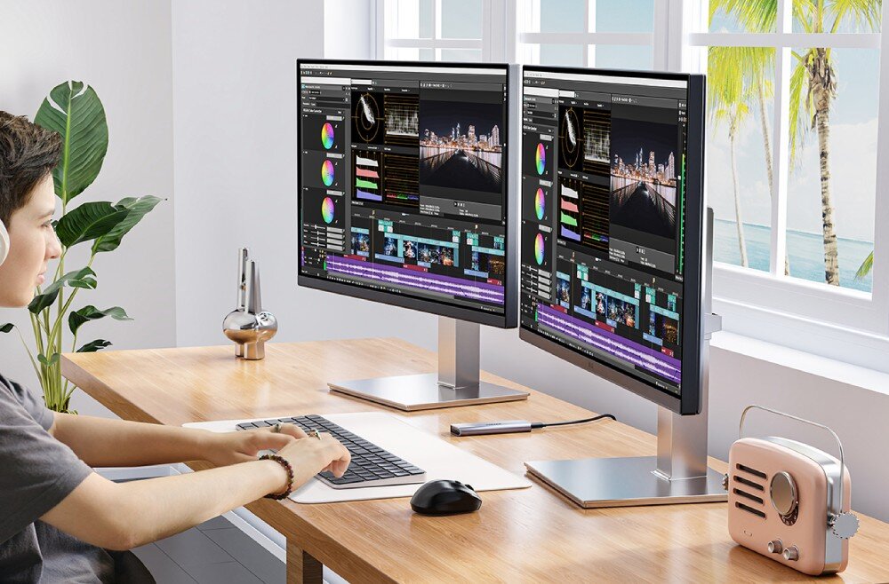 Obudowa dysku UNITEK S1241B01 Osoba siedząca przy drewnianym biurku pracuje na dwóch monitorach z otwartym programem do edycji wideo, obok klawiatury leży podłączona srebrna obudowa na dysk, a w tle widać okno z widokiem na palmy