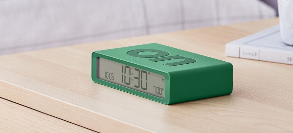 Budzik LEXON Flip Classic zielony, dwustronność, prostota, obrót, wygoda, Budzik silikonowy zielony z napisem off na górze, umieszczony na stoliku, dekoracja kwiatowa obok.