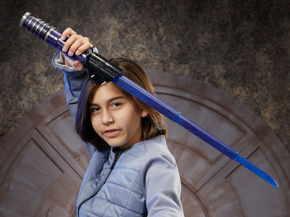 Osoba trzyma niebieski miecz świetlny z czarną rękojeścią. W tle widoczna jest ściana z okrągłym wzorem.