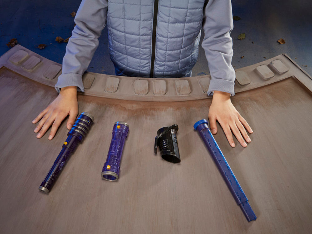 Osoba w szarej kurtce stoi przy stole, na którym leżą części miecza świetlnego w kolorze niebieskim i czarnym. Na stole widoczne są cztery elementy, w tym rękojeść i ostrze.