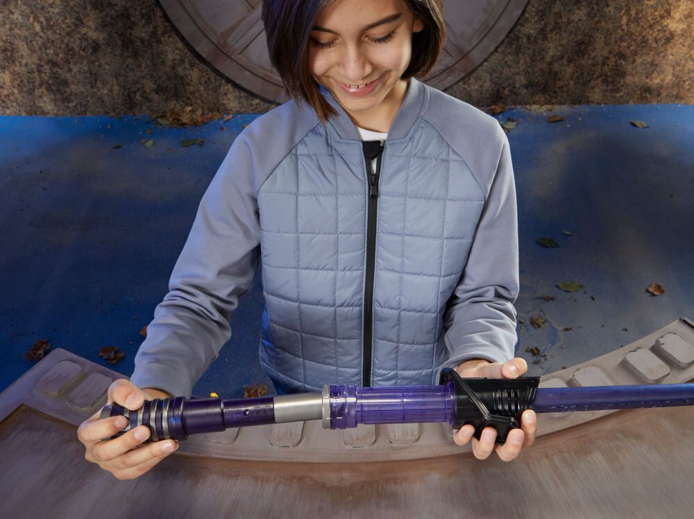 Osoba trzyma miecz świetlny o fioletowym ostrzu i czarnej rękojeści. W tle widać okrągłe drzwi i niebieską podłogę z liśćmi.