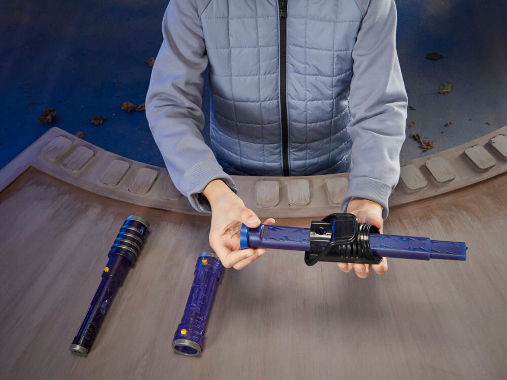 Osoba trzyma niebieski miecz świetlny z czarnymi elementami. Na stole obok leżą dwa podobne miecze świetlne.