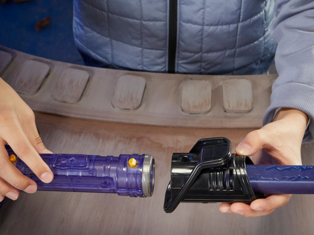 Osoba trzyma dwa elementy miecza świetlnego, jeden z fioletowym uchwytem, drugi z czarnym. Tło jest rozmyte, z widocznymi elementami przypominającymi panel.