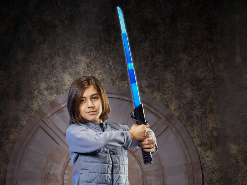 Osoba trzymająca niebieski miecz świetlny na tle okrągłych drzwi. Miecz ma czarną rękojeść i świecące ostrze.