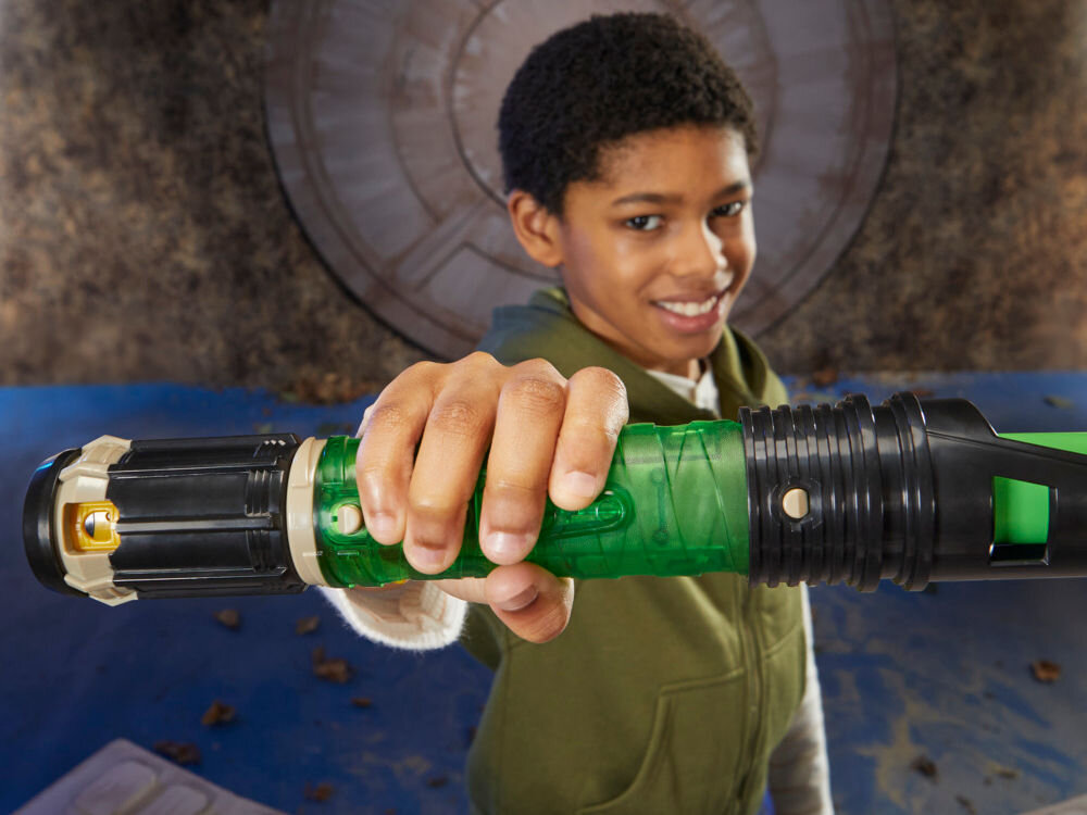 Miecz świetlny HASBRO Star Wars Luke Skywalker F99685X0 Zbliżenie na rękojeść miecza świetlnego Hasbro Lightsaber Forge trzymaną poziomo przez uśmiechniętego chłopca; przezroczysta obudowa odsłania zielony kryształ.