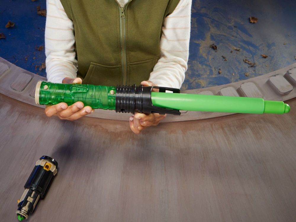 Miecz świetlny HASBRO Star Wars Luke Skywalker F99685X0 Chłopiec w oliwkowej kamizelce trzymający w jednej ręce rozłożony zielony miecz świetlny z serii Lightsaber Forge na tle dekoracyjnej tarczy.