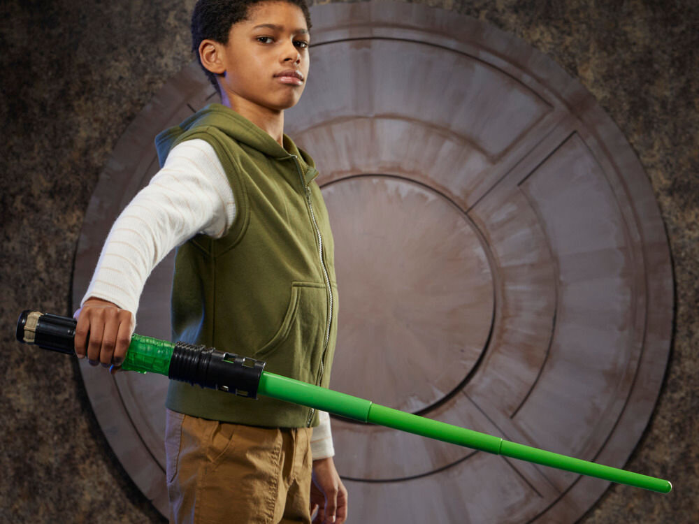 Miecz świetlny HASBRO Star Wars Luke Skywalker F99685X0 Dziecko składa zielony miecz świetlny, łącząc półprzezroczystą obudowę z czarnym emiterem i ostrzem; obok na stole leży gotowa rękojeść.