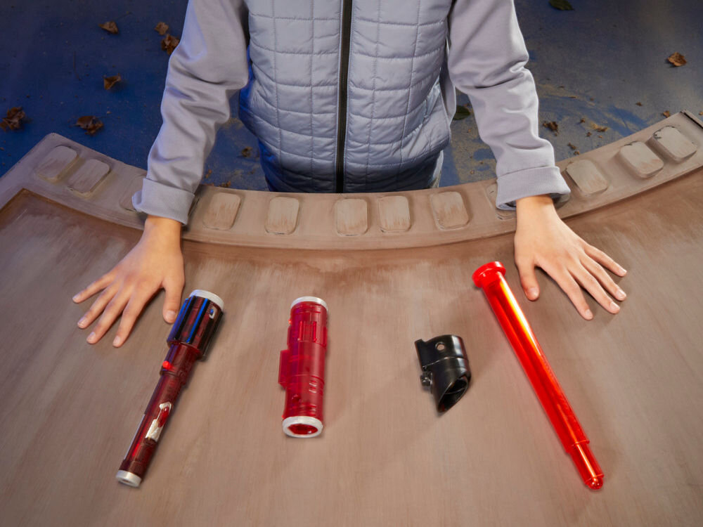 Na drewnianym stole leżą części miecza świetlnego w kolorach czerwonym i czarnym. Osoba w szarej kurtce trzyma ręce na stole obok tych elementów.