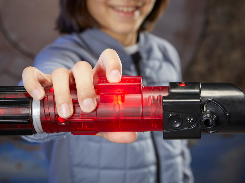 Osoba trzyma miecz świetlny z czerwonym, przezroczystym uchwytem i czarnymi elementami. W tle widoczna jest rozmyta postać w jasnej kurtce.