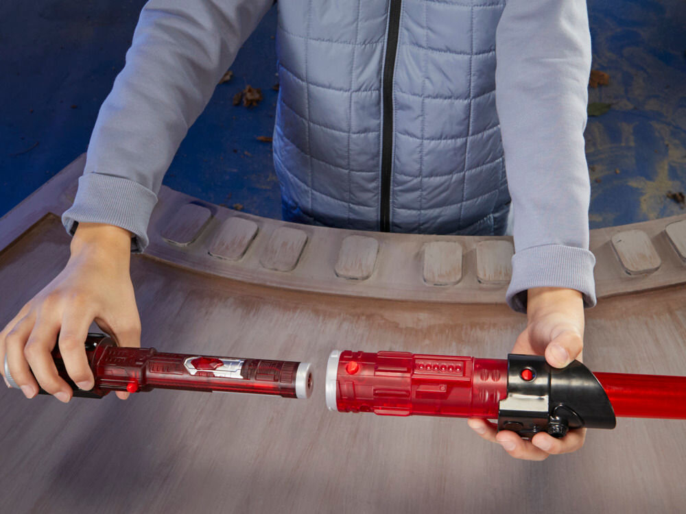 Osoba trzyma czerwony miecz świetlny, który jest rozkładany na dwie części. Miecz ma czarną rękojeść z czerwonym przyciskiem.