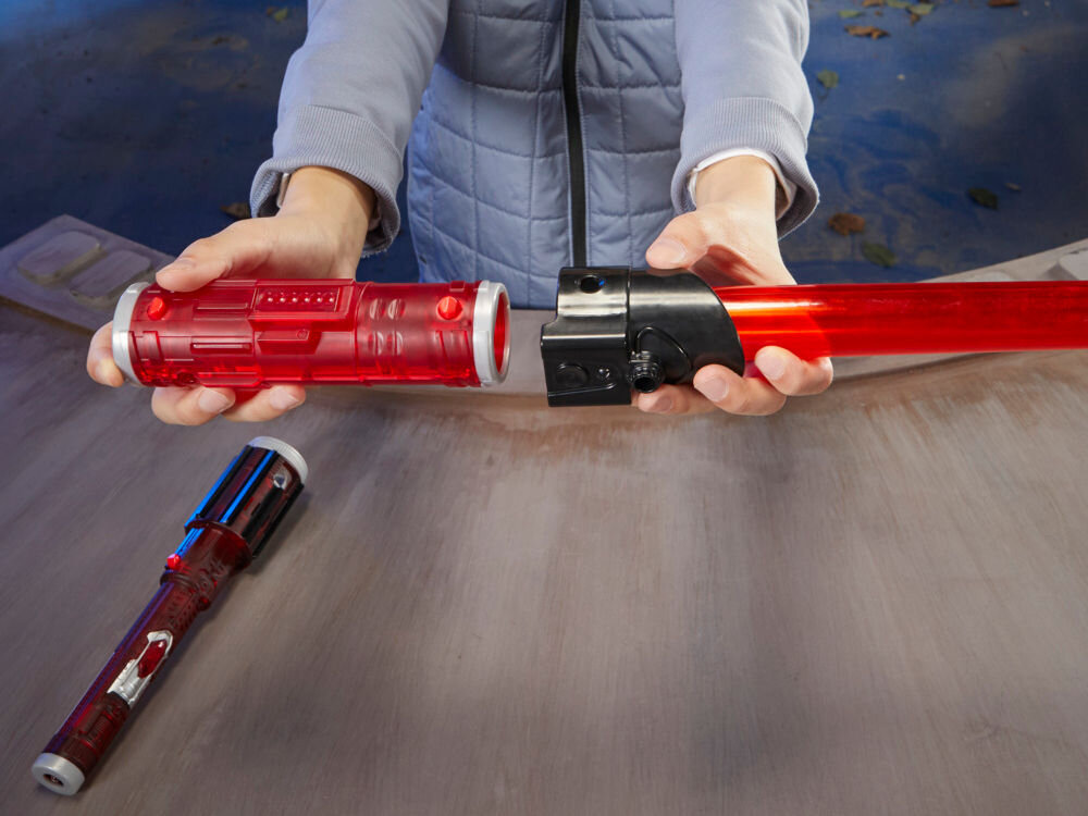 Osoba trzyma czerwony miecz świetlny z czarną rękojeścią na drewnianym stole. Obok leży drugi, podobny miecz świetlny.