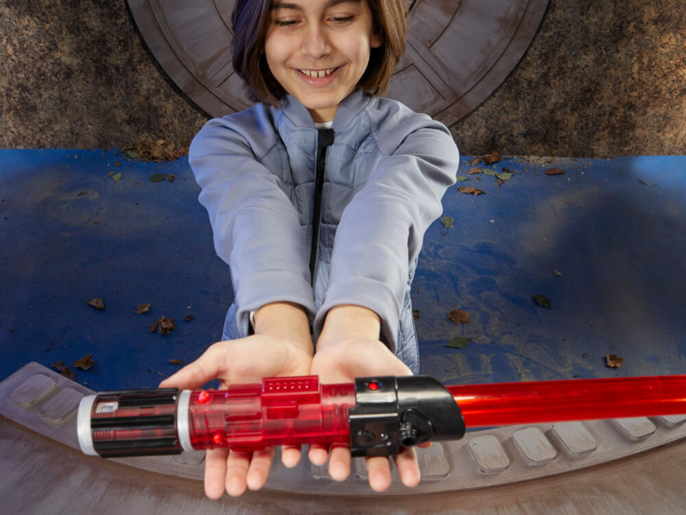 Osoba trzyma czerwony miecz świetlny z czarnymi i srebrnymi elementami na rękojeści. W tle widoczna jest niebieska podłoga z liśćmi i okrągła struktura.