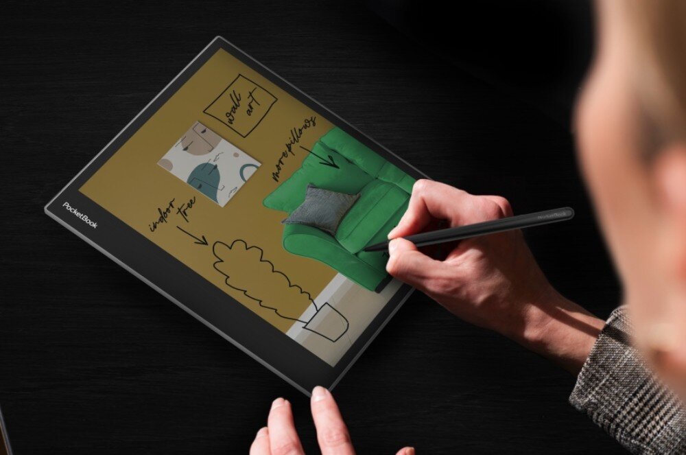Czytnik e-booków POCKETBOOK InkPad Eo Widok z góry na czytnik leżący na czarnym stole. Użytkownik rysuje rysikiem po zdjęciu przedstawiającym wnętrze z zielonym fotelem, rośliną i obrazami. Na zdjęciu znajdują się także odręczne notatki, rysik, rozmiar, notatki