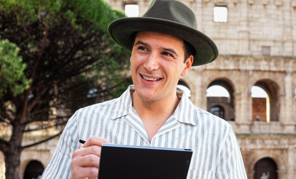 Czytnik e-booków POCKETBOOK InkPad Eo Uśmiechnięty mężczyzna w kapeluszu i pasiasta koszuli trzyma czytnik i rysik, stojąc na tle Koloseum. Spogląda z zadowoleniem poza kadr, aparat, zdjęcia