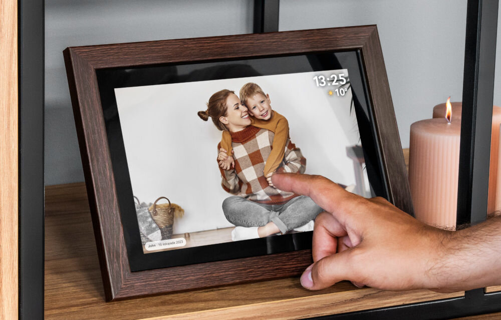 Ramka cyfrowa KRUGER&MATZ KM1102-15 Frameo 15.6 cala Brązowy prezentacja ramki ze zdjęciem i komentarzem do niej na regale palec dotykający ramkę obok świeczki 15 calowy ekran rozdzielczość IPS komfortowe przeglądanie