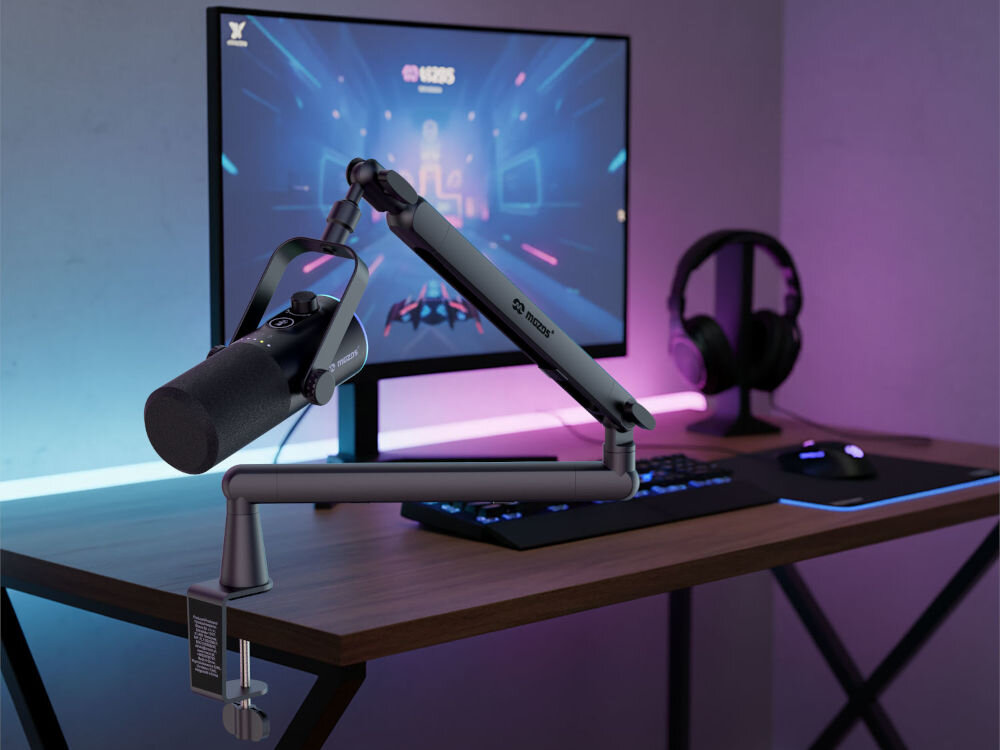 Zestaw mikrofonowy Mozos TAM8 zamontowany na biurku przy stanowisku gamingowym. Do mikrofonu przymocowane jest metalowe ramię z regulacją, a w tle widać monitor z dynamiczną grafiką, klawiaturę, mysz oraz słuchawki na uchwycie