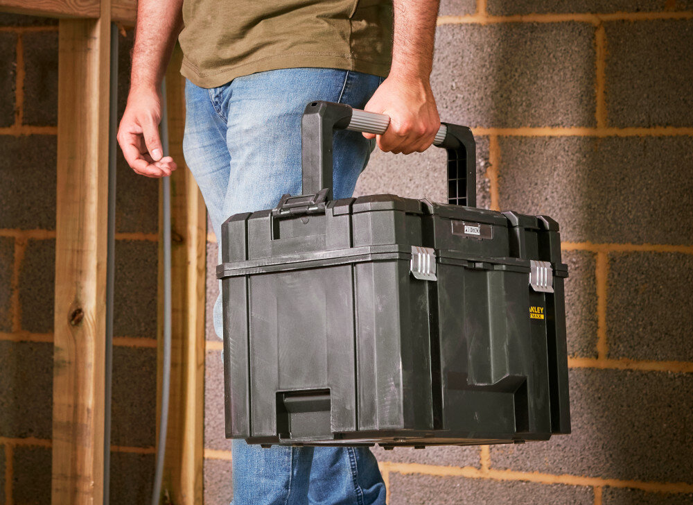 Skrzynka narzędziowa STANLEY Fatmax TSTAK FMST1-75796 pomieszczenie osoba trzyma w dłoni skrzynkę w tle murowana ściana długa aluminiowa rączka pewny chwyt komfort podczas przenoszenia waga 3,3 kg aluminiowy uchwyt trwałość wygoda