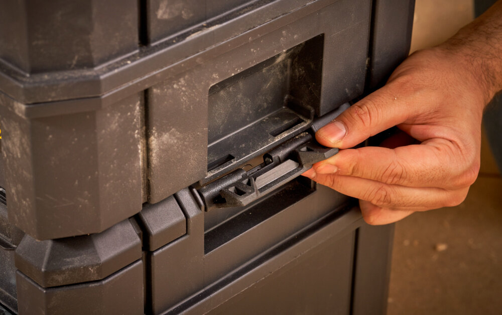 Skrzynka narzędziowa STANLEY Fatmax TSTAK FMST1-75796 pomieszczenie skrzynia ustawiona na innej skrzyni osoba trzyma zatrzaski w dłoniach część systemów STANLEY FATMAX PRO-STACK™ i TSTAK łączenie z innymi modułami dopasowane zestawy boczne zatrzaski szybkie i stabilne łączenie oraz rozłączanie skrzyń wygodne mobilne stanowisko pracy