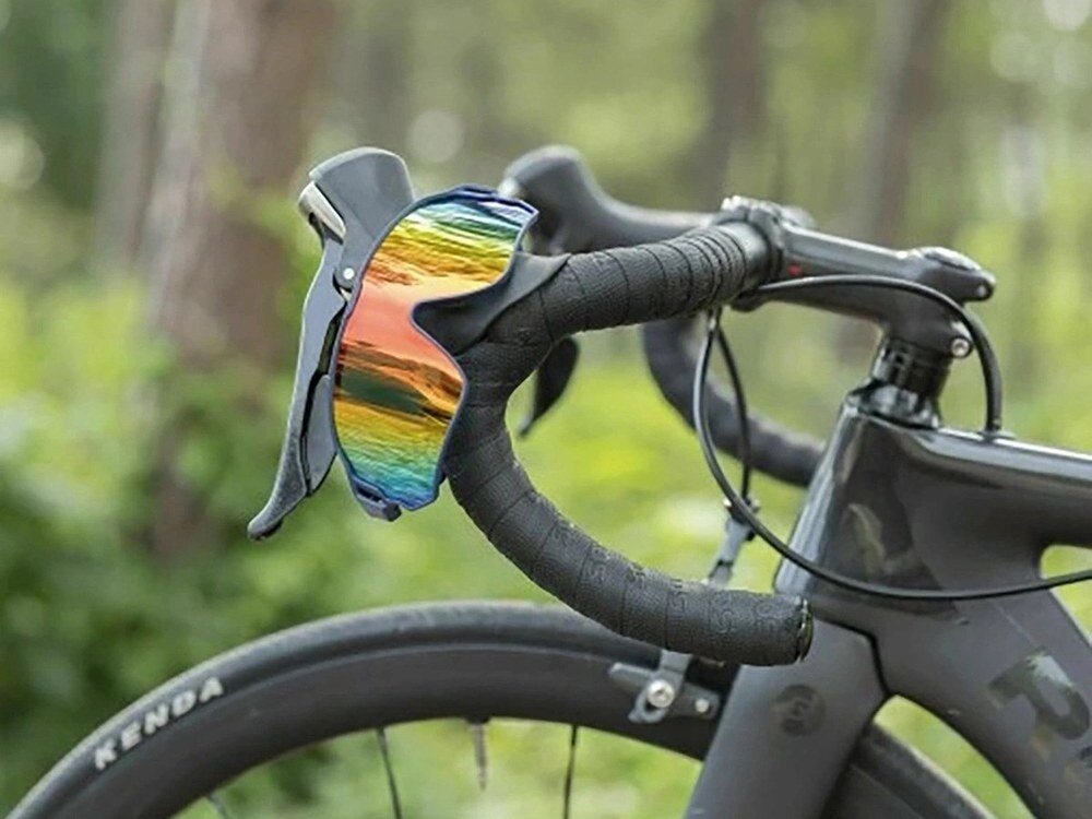 Okulary rowerowe z kolorowymi, lustrzanymi soczewkami zawieszone na kierownicy roweru. W tle widoczna jest rozmyta zieleń lasu.