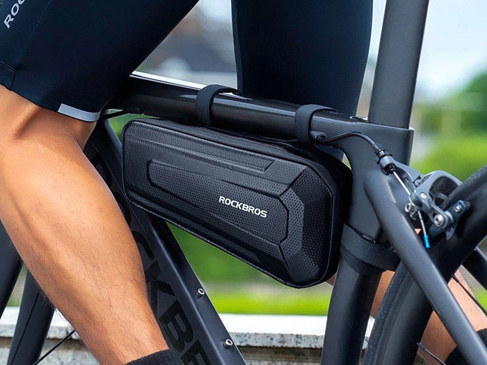 Na zdjęciu widać czarną torbę rowerową zamocowaną na ramę roweru z napisem \'ROCKBROS\'. Torba jest przymocowana za pomocą pasków i znajduje się w pobliżu pedałów roweru.