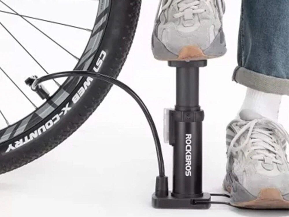 Osoba używa nożnej pompki rowerowej z manometrem, na której widnieje napis \'ROCKBROS\'. Obok znajduje się koło rowerowe z widocznym napisem \'CST X-COUNTRY\'.