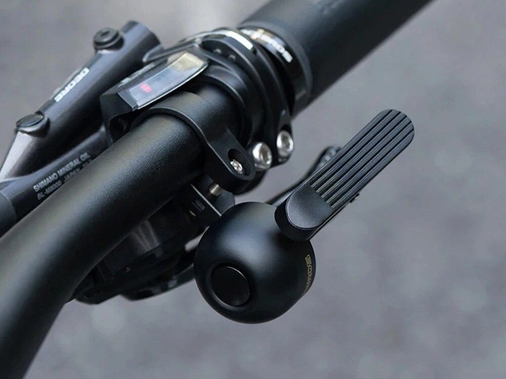 Czarny dzwonek rowerowy zamontowany na kierownicy roweru. Obok widoczny jest fragment manetki z napisem \'SHIMANO\'.