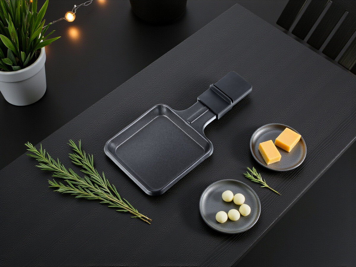 Grill elektryczny MELISSA 16300027, patelnie, bezpieczeństwo, uchwyty, kontrola, Sama patelnia raclette z zestawu MELISSA 16300027 na czarnym stole, obok gałązka rozmarynu i kawałki sera na talerzykach.