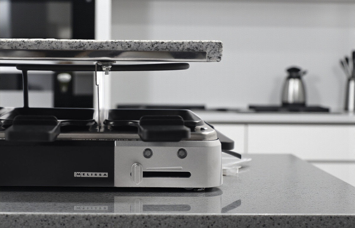 Grill elektryczny MELISSA 16300027, czystość, prostota, mycie, przechowywanie, Zbliżenie na bok grilla MELISSA 16300027 z widoczną kamienną płytą i wysuniętymi patelniami raclette, w tle biała kuchnia.