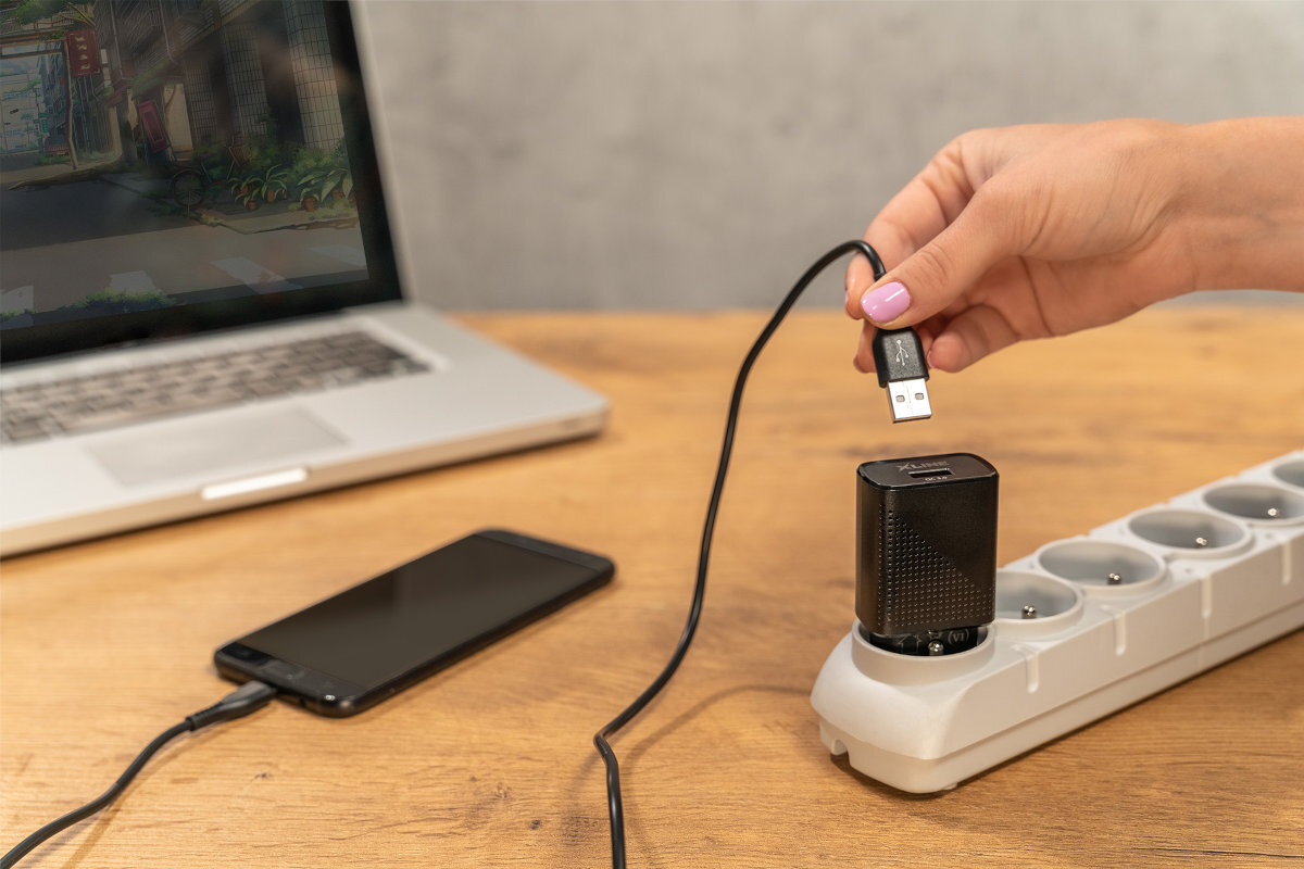 Osoba trzyma kabel USB, który jest podłączony do czarnej ładowarki sieciowej umieszczonej w listwie zasilającej. Obok na stole znajduje się smartfon i otwarty laptop.