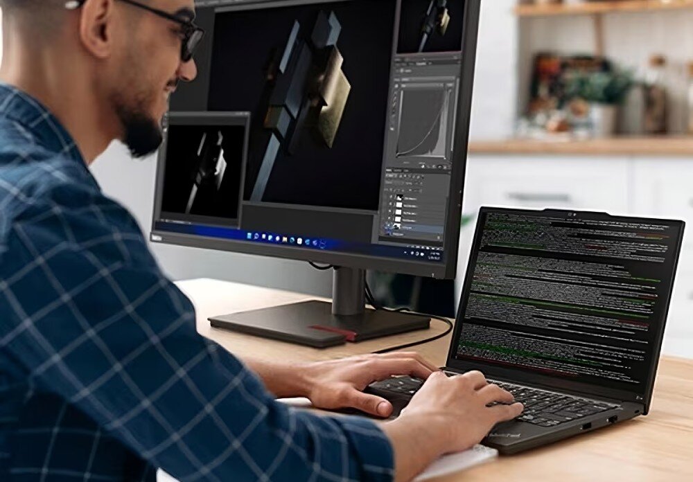 Programista w okularach pracujący na laptopie ThinkPad przy domowym stanowisku, obok monitor z oprogramowaniem do modelowania 3D