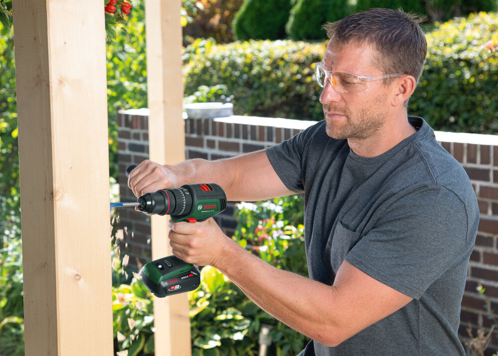 Wiertarka udarowa BOSCH AdvancedImpact 18V-80 06039E2101 podwórko ogród osoba pracuje przy drewnianym elemencie w tle płot z cegły kwiaty krzewy drzewa moc precyzja wielofunkcyjność