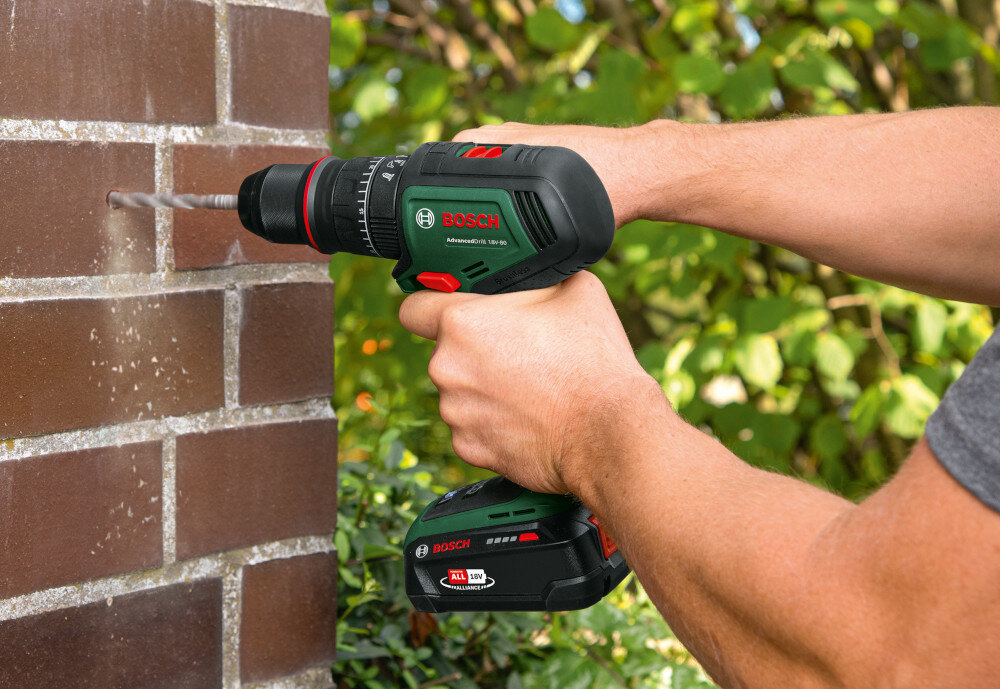 Wiertarka udarowa BOSCH AdvancedImpact 18V-80 06039E2101 ceglana ściana osoba wierci otwór w ścianie w tle liście rośliny maksymalny moment obrotowy na poziomie do 82 Nm regulacja prędkości obrotowej dostosowanie siły i tempa wiercenia lub wkręcania do materiału efektywność skraca czas pracy 22-stopniowa regulacja momentu pełna kontrola nad procesem pracy