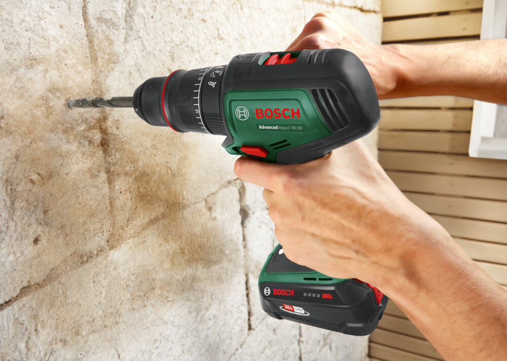 Wiertarka udarowa BOSCH AdvancedImpact 18V-80 06039E2101 ściana z dużej cegły w tle drewniana ściana osoba wierci otwór w ceglanej ścianie silnik bezszczotkowy metalowy uchwyt wiertarskim 13 mm wytrzymałość długa żywotność precyzyjne działanie szybkozaciskowy uchwyt
