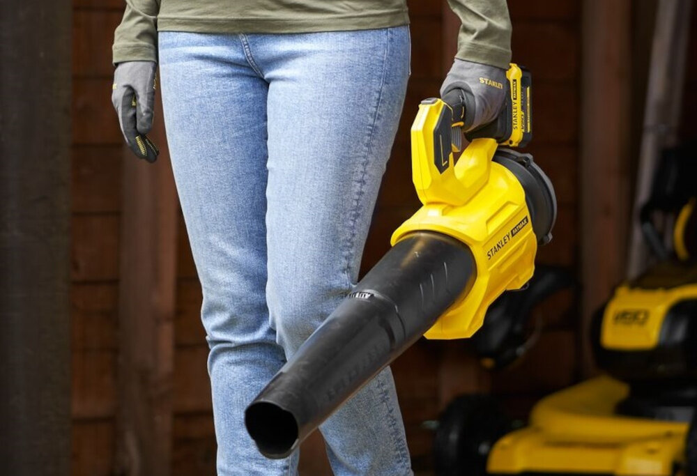 Dmuchawa do liści STANLEY SFMCBL730B-XJ akumulatorowa rozmazane tło osoba niesie dmuchawę w dłoni lekka waga 3,4 kg stabilna wygodna w użytkowaniu ergonomiczny uchwyt pewny chwyt minimalizuje zmęczenie dłoni praktyczny hak do zawieszenia kompatybilny z systemem ściennym STANLEY® Track ochrona przed uszkodzeniami oszczędność miejsca