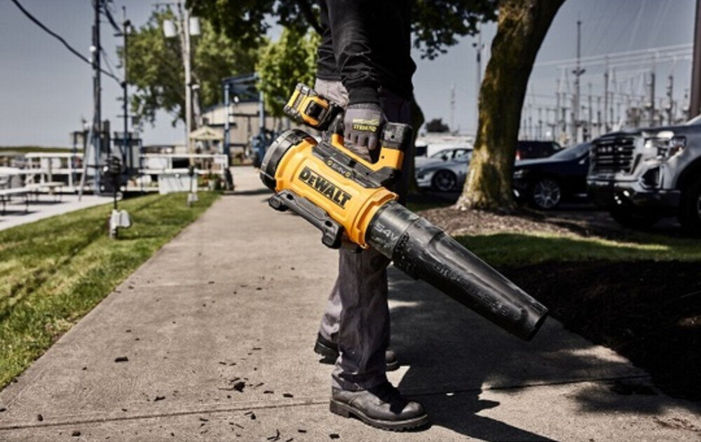 Dmuchawa do liści akumulatorowa DEWALT DCMBL777X1-QW (z akumulatorem) osoba trzyma dmuchawę w dłoni zdmuchuje liście w tle samochody niebo sprzątanie porządek mobilność