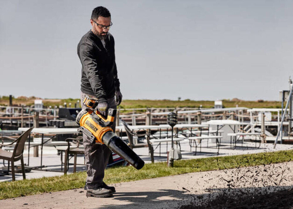 Dmuchawa do liści akumulatorowa DEWALT DCMBL777X1-QW (z akumulatorem) osoba z dmuchawą w dłoni zdmuchuje liście z drogi w tle meble ogrodowe trawnik prędkość powietrza do 258 km/h przepływ do 21,5 m³/min sprawne usuwanie liści i brudu nawet z trudno dostępnych miejsc