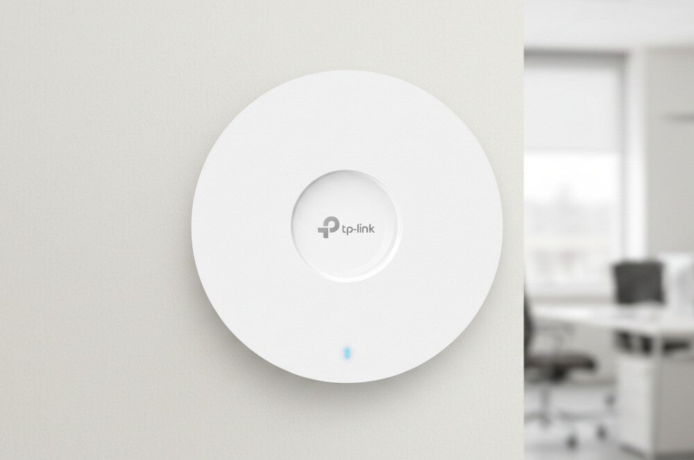 Na ścianie znajduje się biały, okrągły punkt dostępu z logo \'tp-link\' na środku. W tle widoczne jest rozmyte wnętrze biura.