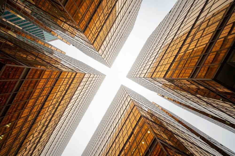Widok z dołu na cztery szklane wieżowce zbiegające się ku środkowi kadru, z jasnym niebem pośrodku