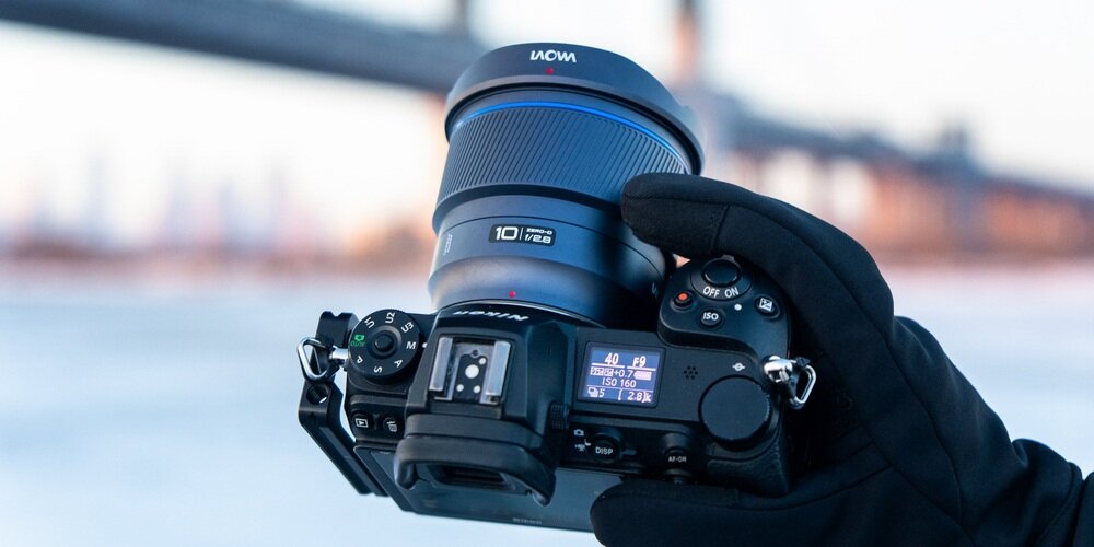Aparat fotograficzny trzymany w rękawiczkach z obiektywem LAOWA, w tle rozmyty most nad wodą