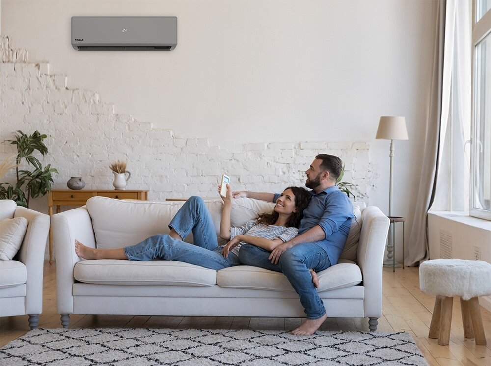 Na ścianie wisi klimatyzator z logo \'FINLUX\'. Na kanapie siedzi para, a obok znajduje się lampa stojąca i stolik z dekoracją.