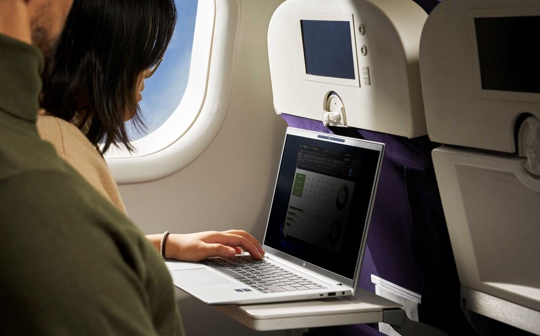 Kobieta pracuje na srebrnym laptopie HP EliteBook 845 podczas lotu samolotem; laptop stoi na stoliku przy oknie, na ekranie wyświetlony arkusz kalkulacyjny, w tle siedzenia i ekrany pokładowe. - Stabilne połączenie Wi-Fi 6E