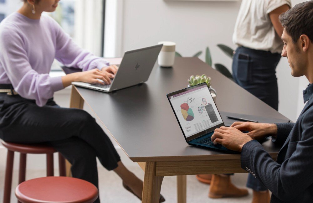 Laptop HP EliteBook 630 G11 Dwie osoby pracujące na laptopach przy stole w przestrzeni biurowej, jedna osoba stoi w tle, opis