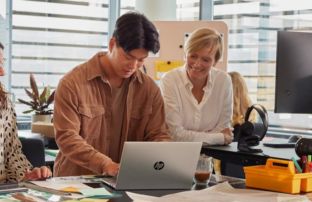 Laptop HP ProBook 445 G11 - grupa osób pracujących przy biurku w biurze. Mężczyzna w brązowej koszuli korzysta z laptopa, a kobieta w białej koszuli uśmiecha się i obserwuje ekran